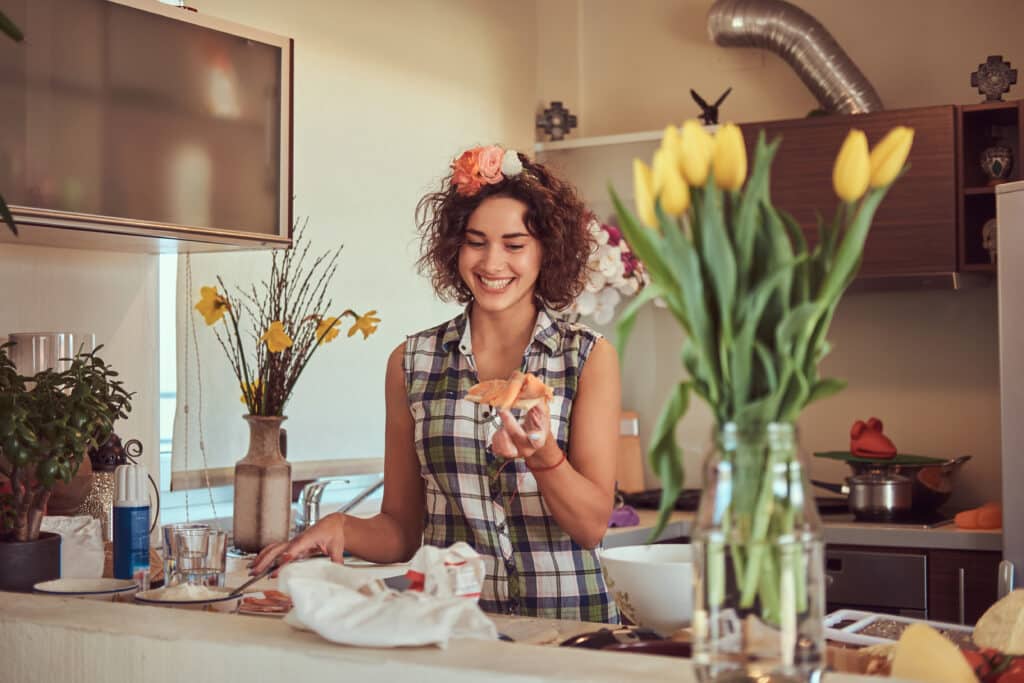 Smiling woman with curly hair and flower crown holding homemade pizza dough in a warm, plant-filled kitchen