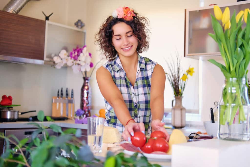 Woman with curly hair and a flower crown gently arranging tomatoes in a bright kitchen surrounded by plants and cheese